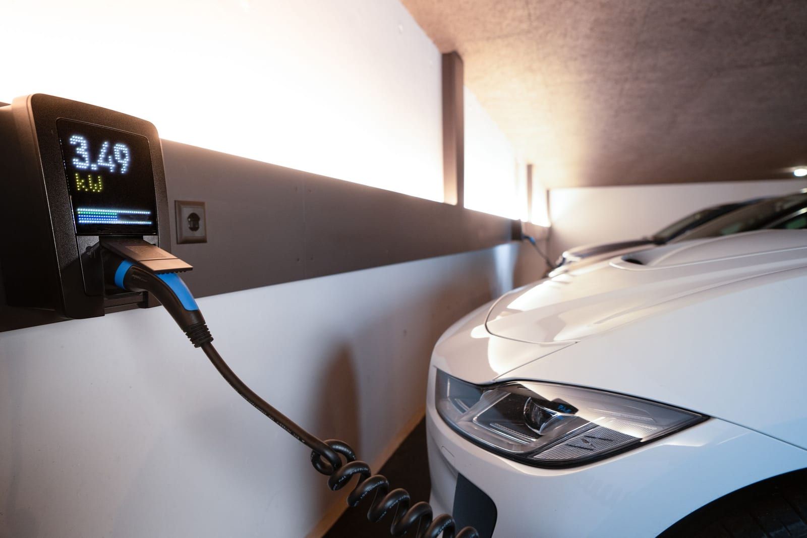 Wallbox mit Display an einer Garagenwand, daneben ein ladendes Elektroauto